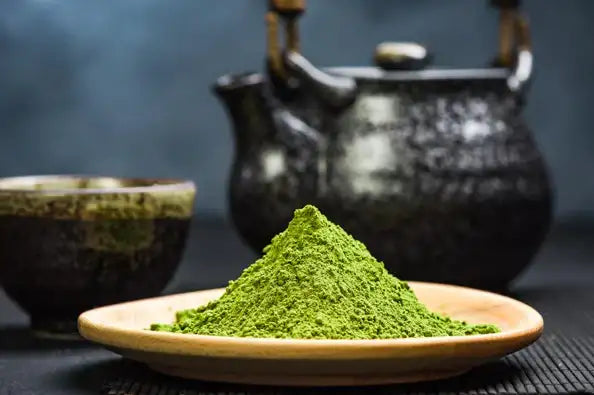 Matcha-Grüntee-Pulver auf einem Holzteller mit schwarzem Keramikteekessel und Schalen im Hintergrund.