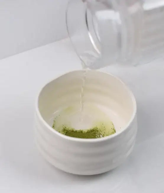 Weiße Keramikschüssel mit grünem Pulver und klarer Flüssigkeit, die hineingegossen wird